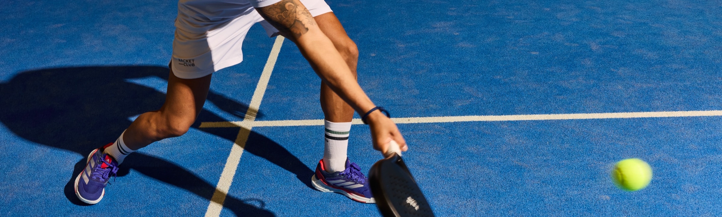 Padelspiller i udfald på en blå bane med battet strakt mod bolden og skarpe skygger