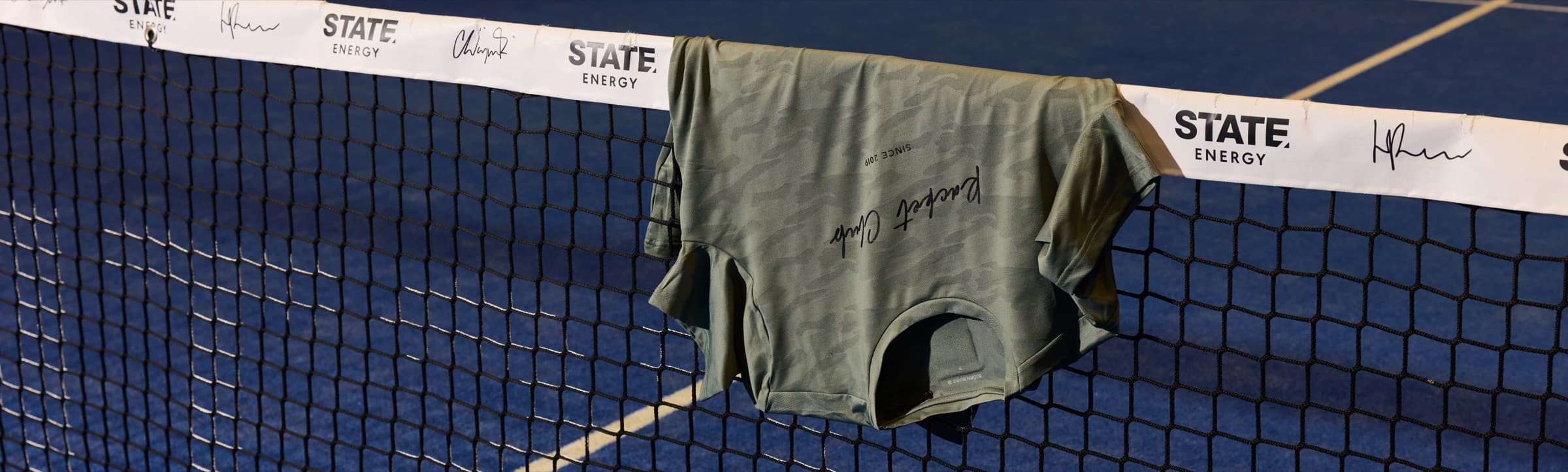 Racket Club-trøje draperet over padelnettet på en blå bane